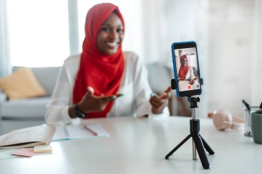 Evde vlog için içerik yakalayan pozitif siyah islami kadın, tripodla akıllı telefondan video kaydetmek, cihaz kamerasına konuşmak ve işaret etmek, seçici odaklanma