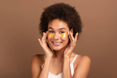 Kıvırcık saçlı, göz bandı takan, prosedürlerden hoşlanan kahverengi arka planda, stüdyoda izole edilmiş mutlu, genç, melez bir ırk kadını. Cilt bakımı, doğal güzellik, spa tedavisi ve covid-19 'a bağlı yaşam tarzı.