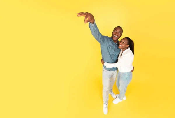 Look At This. Happy African American Spouses Advertising Something Holding Invisible Object In Fingers Posing Standing Near Copy Space Over Yellow Background. Studio Shot