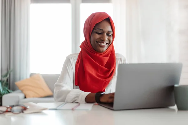 Remote job. Happy islamic woman in red hijab working on laptop, typing on keyboard and looking at computer screen, sitting at workplace at home office