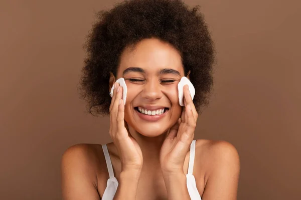 Happy young mixed race lady with curly hair removes makeup from face with cotton pad isolated on brown background, close up. Lifestyle, treatment of problem skin, natural beauty care, spa therapy