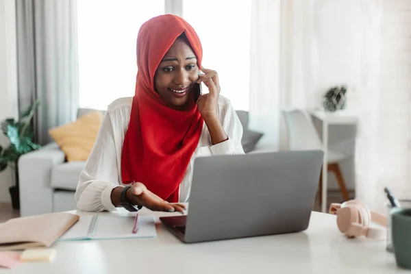 Problem concept. Frustrated black muslim woman talking on cellphone, sitting at desk and using laptop, having problem with internet connection, calling customer support hotline