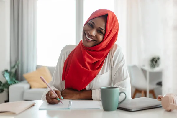 Note taking concept. Happy black muslim woman sitting at desk with laptop and writing letter in paper notebook, smiling at camera. Excited black islamic lady planning her day