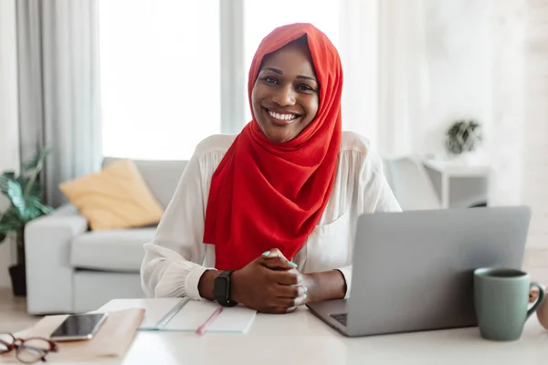 Portrait of black muslim freelancer woman in hijab sitting at table with laptop and smiling at camera, islamic lady working remotely on computer at home office, copy space