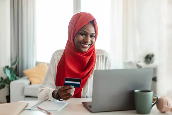 Cashless payment. Happy black muslim woman in hijab holding credit card and using laptop, shopping online from home. Lady using electronic money to buy goods in web store