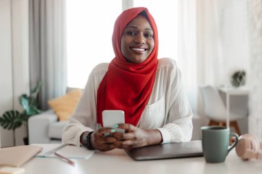 Afro-Amerikalı, tesettürlü, akıllı telefon kullanan, masada oturan, kameraya gülümseyen, arkadaşlarıyla mesajlaşan ya da sosyal ağlara göz atan, boş alan arayan Afro-Amerikan kadın.