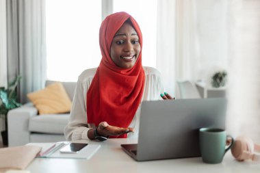 Afro-Amerikan Müslüman kadın video görüşmesi yapıyor, dizüstü bilgisayara bakıyor ve el kol hareketi yapıyor, oturma odasında iş yerinde oturuyor. İş ve ev ofisinde buluşma, covid-19 karantinası sırasında sosyal mesafeler.
