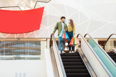 Kara Cuma indirimi. Alışveriş maskesiyle alışveriş yapan, alışveriş merkezinde alışveriş yapan merdivenlerde duran alışveriş torbaları taşıyan bir çift. Tüketim ve Alışverişcilik. Tam Uzunluk