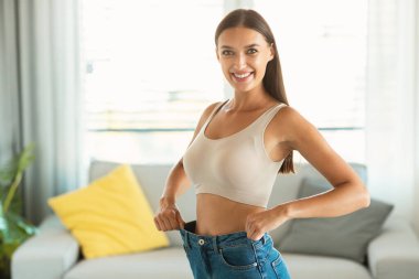 Sıska kadın, büyük beden kot pantolon giyiyor. Kilo verdikten sonra evde poz veriyor, kameraya gülümsüyor. Diyet Sonuçları ve Başarılı Zayıflama Konsepti