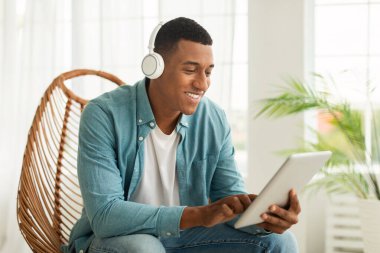 Kablosuz kulaklıklı, gülümseyen Afro-Amerikan bir adam. Sohbet ediyor, arıyor, internette salonun içindeki tablette sörf yapıyor. Evde çalış ve çalış, sosyal uzaklık ve aygıtla yeni normal