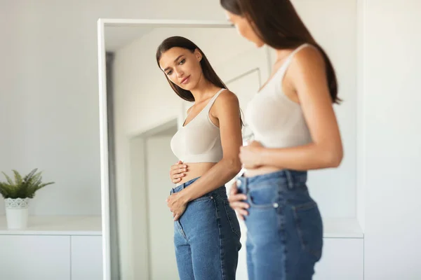 Young Woman Touching Flat Belly Expecting Baby And Enjoying First Months Of Pregnancy Posing Looking At Her Reflection In Mirror Wearing Skinny Jeans Standing At Home. Selective Focus