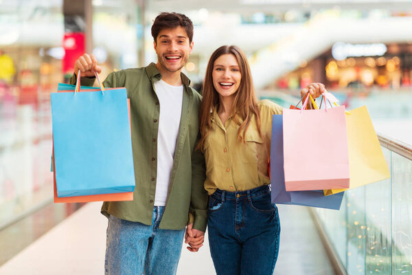 Предложение о продаже. Счастливые супруги держат бумажные пакеты Shopper Shopper вместе позируя в гипермаркете, улыбаясь глядя на камеру. Морские скидки и черная пятница