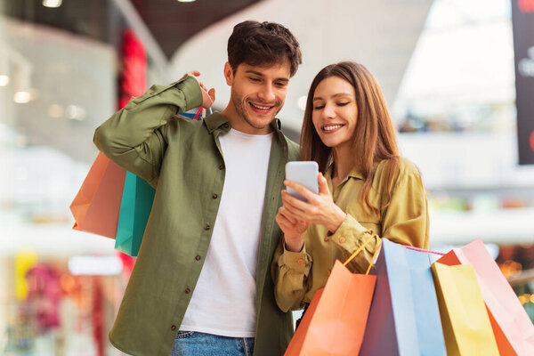 Веселые покупатели пара делает покупки в Интернете через смартфон приложение Позирование Холдинг красочные сумки Shopper в современном гипермаркете. Концепция продаж и электронной коммерции