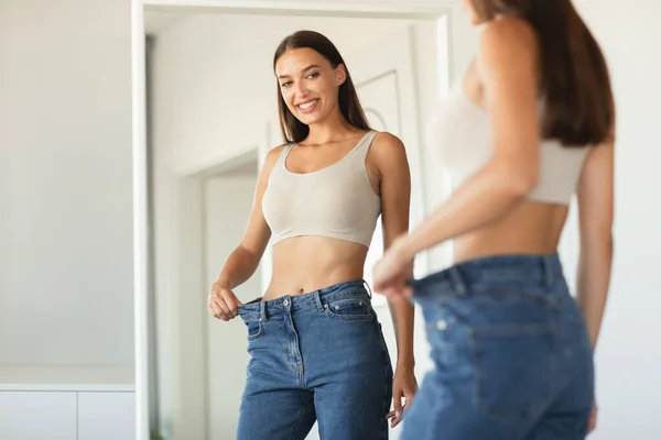 痩身コンセプト 自宅で鏡の近くに立っているサイズのポーズを比較すると 大きな減量後のジーンズを身に着けて興奮したスリム女性 選択的フォーカス 切り抜きショット — ストック写真