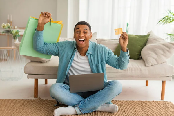兴奋的千年非洲裔美国男性 带着购物袋和信用卡在笔记本电脑上购物 享受在客厅里的销售 支付检疫期间的定单 — 图库照片