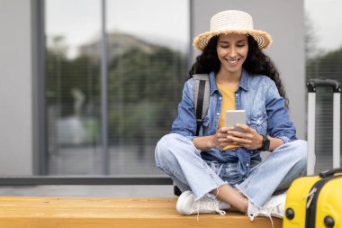 Uzun saçlı neşeli bir genç bayan havaalanının yanındaki bankta oturuyor, modern cep telefonu kullanıyor ve gülümsüyor, bayan yolcu rezervasyonu, otel veya biletleri mobil uygulama üzerinden, kopyalama alanı kullanıyor.