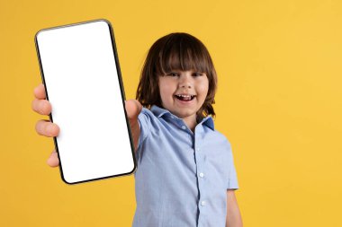 Çocuklar için harika bir uygulama. Küçük mutlu çocuk kameraya boş ekranlı akıllı telefon gösteriyor ve gülümsüyor, turuncu stüdyo arka planı, boş alan