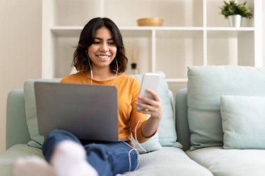 Evde koltukta dinlenirken dizüstü bilgisayar ve akıllı telefon kullanan gülümseyen genç Arap Kadın, arkadaşlarıyla cep telefonunda mesajlaşan veya yeni uygulamalara göz atan Orta Doğulu Kadın, Boşluğu Kopyala