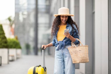 Hasır şapkalı, elinde akıllı telefonuyla havaalanının yanında duran, çanta ve bavul taşıyan seyahat uygulaması kullanan, taksi bekleyen, fotokopi alanı olan çekici, uzun saçlı genç bir kadın.