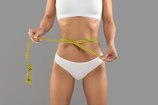 重量損失の概念 テープでウエストを測定下着でスポーティボディを持つ若い女性 ダイエット後の測定をスレンダーフィギュアチェックと認識できない女性 グレーの背景に立って — ストック写真