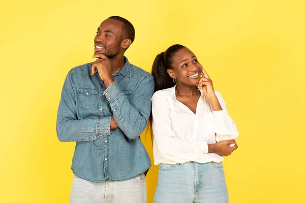 Happy African American Couple Thinking And Dreaming About Something Touching Chin Looking In Different Sides Posing Standing Over Yellow Background. Studio Shot