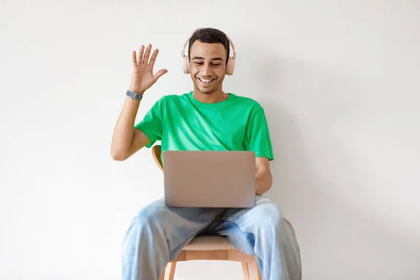 Excited arab man video calling via laptop and waving hello to webcamera, sitting on chair over light wall background. Cheerful guy wearing wireless headphones and smiling