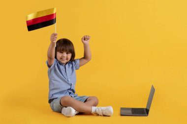 En küçük çocuklar için yabancı kurslar. Almanya bayrağı taşıyan heyecanlı küçük çocuk dizüstü bilgisayarın yanında oturuyor ve gülümsüyor, online eğitimin tadını çıkarıyor, turuncu stüdyo geçmişi, bedava alan.