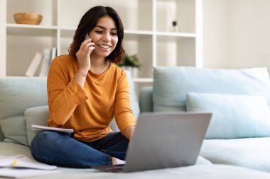 Uzaktan kumanda. Orta Doğulu genç bir kadın evinde cep telefonu ve dizüstü bilgisayar kullanıyor bilgisayarla çalışan ve oturma odasında koltukta otururken cep telefonuyla konuşan gülümseyen bir Arap kadın serbest çalışıyor.