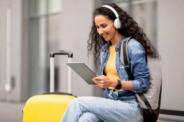 Sırt çantalı, valizli, dijital tablet ve kablosuz kulaklık kullanan, dışarıda bankta oturup internetten film izleyen esmer bir kadın. Seyahat ederken eğlence