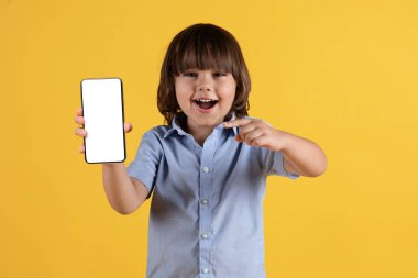 Çocuklar ve aletler. Beyaz ekranlı, gülümseyen, uygulama öneren, turuncu stüdyo geçmişi olan boş bir telefonu işaret eden mutlu küçük çocuk.
