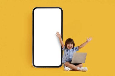 E öğrenme konsepti. Modern aletlerle online eğitimden hoşlanan sevimli küçük çocuk, dizüstü bilgisayar taşıyor, beyaz ekranlı büyük akıllı telefonun yanında oturuyor, turuncu stüdyo arka planı, boş alan