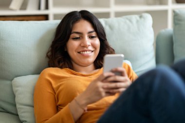 Evde dinlenirken cep telefonuyla mesajlaşan gülümseyen genç Arap kadın, oturma odasındaki koltukta dinlenen mutlu Orta Doğulu kadın ve çevrimiçi iletişim için akıllı telefonu kullanan,