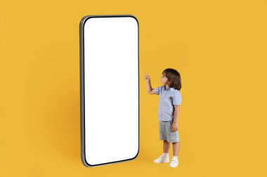 Heyecan verici modern teknolojiler. Beyaz ekranlı büyük boş akıllı telefonu işaret eden sevimli küçük çocuk, cihazdaki içeriği seçiyor, turuncu stüdyo arka planı, serbest alan