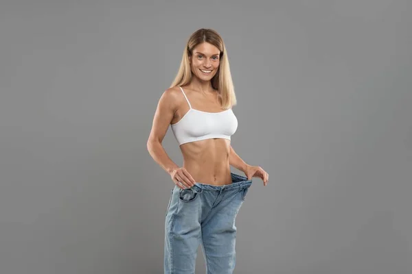 Weight Loss Result. Happy Young Woman Posing In Oversized Jeans, Slender Female Wearing Big Clothes Standing On Grey Studio Background, Comparing Sizes After Successful Diet, Copy Space