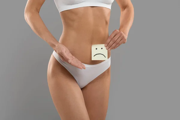 Health Problems Concept. Young Woman In Underwear Holding Paper Card With Drawing Unhappy Face Near Belly, Unrecognizable Female Standing Isolated Over Grey Studio Background, Cropped