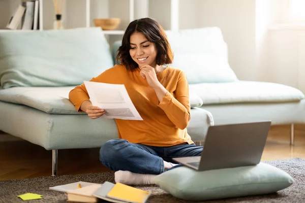 Arab Female Freelancer Working With Papers And Laptop While Sitting On Floor At Home, Smiling Young Middle Eastern Woman Checking Financial Reports And Using Computer, Enjoying Remote Work