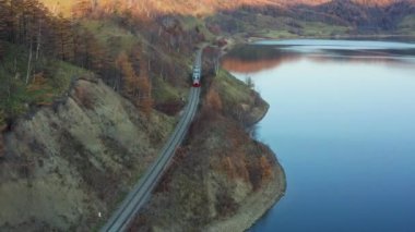 Tren, çam ağaçlarıyla dolu bir dağ ormanı gölünde turkuaz su boyunca yol alır. Trenin, gölün ve yeşil ormanların havadan görünüşü. Dağ ormanları arasındaki göl manzarası Yolcu treni kristal berrak su üzerinden geçiyor