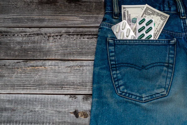 Kot pantolonun arka cebinde bir paket hap. Ahşap bir masanın arka planında pantolon cebinde dolar banknotları. Mevsimsel grip virüsü konsepti.