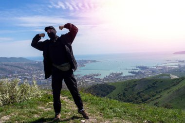 Başlık ve kar maskeli bir adam, siyah bir yağmurluk ve yürüyüş botu giymiş bir adam şehrin arka planına karşı dağlarda poz veriyor. Yeşil tepeler ve liman. Novorossiysk, Krasnodar Bölgesi, Rusya.