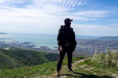 Başlık ve kar maskeli bir adam, siyah bir yağmurluk ve yürüyüş botu giymiş bir adam şehrin arka planına karşı dağlarda poz veriyor. Yeşil tepeler ve liman. Novorossiysk, Krasnodar Bölgesi, Rusya.