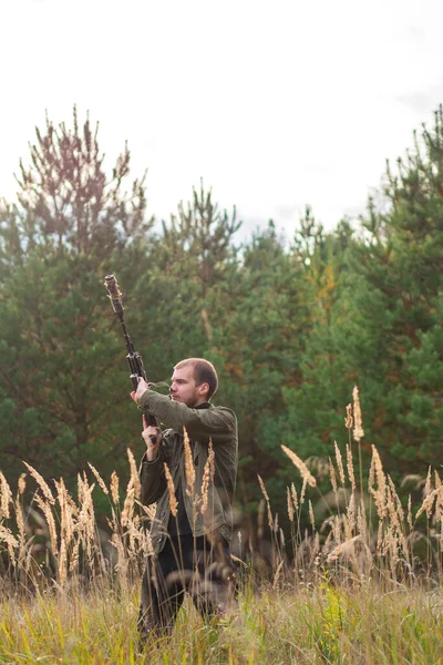 A man in civilian clothes reloads an automatic rifle. A man with a gun in an autumn field against the background of a pine forest. Survival in the wild, hunting.