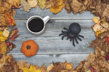 Gri ahşap bir masada, balkabağı oyuncağının yanında siyah bir örümceğin olduğu beyaz bir fincan kahve, sonbahar sarısı yaprakların arka planına karşı. Cadılar Bayramı ve araknofobi kavramı.