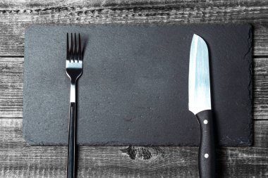 Ahşap bir masa arkasında servis çatalı ve bıçak olan boş bir siyah tahta kesme tahtası. Kopyalama alanı eklenecek yerdir.