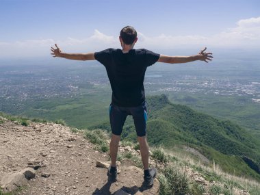 Siyah tişörtlü bir adam şehrin arka planına karşı yüksek bir dağda duruyor. Arkasından sarılan, geniş kollu bir adamın fotoğrafı. Pyatigorsk, Kafkasya 'daki Beshtau Dağı