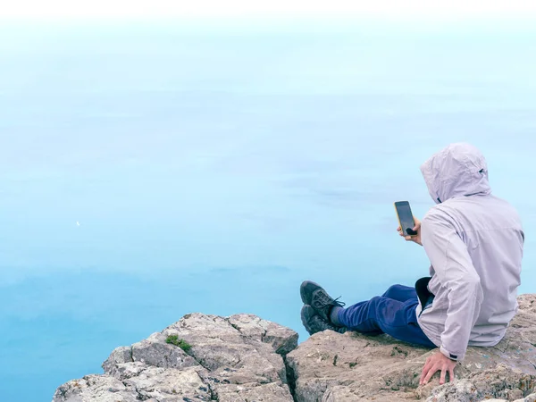 Man in a gray windbreaker sits on a sheer cliff above the serene black sea. Man shares his impressions on a hike with friends via video link.