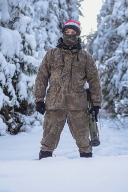 A military man in camouflage is holding a rocket launcher in the outdoors. A soldier with a gun in the winter forest. A male in a red Christmas hat.