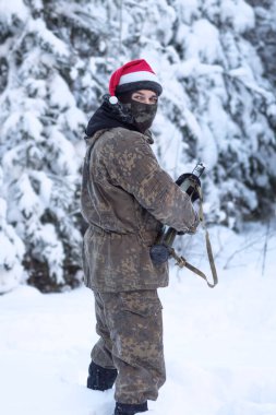 A military man in camouflage is holding a rocket launcher in the outdoors. A soldier with a gun in the winter forest. A male in a red Christmas hat.