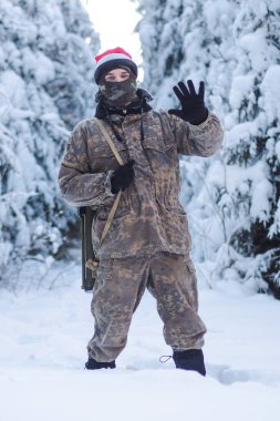 A military man in camouflage is holding a rocket launcher in the outdoors. A soldier with a gun in the winter forest. A male in a red Christmas hat.