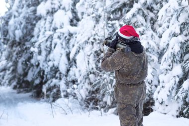A military man in camouflage is holding a rocket launcher in the outdoors. A soldier with a gun in the winter forest. A male in a red Christmas hat.
