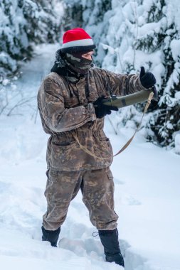 A military man in camouflage is holding a rocket launcher in the outdoors. A soldier with a gun in the winter forest. A male in a red Christmas hat.
